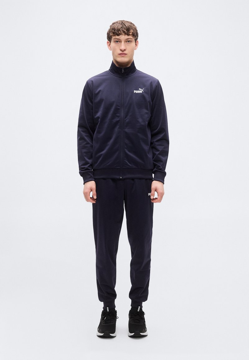 Σκούρο μπλε φόρμα με ζακέτα με φερμουάρ και παντελόνι με ελαστική μέση. Διαθέτει λογότυπο στο στήθος και αθλητικά παπούτσια. Ομαλό, ελαφρύ υλικό.