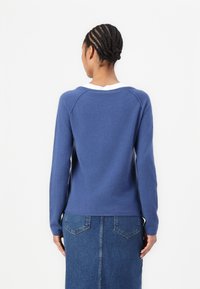 Μπλε πλεκτό πουλόβερ με μακριά μανίκια, στρογγυλή λαιμόκοψη και ριγιέ μανσέτες. Συνδυάζεται με τζιν φούστα που διαθέτει δύο πίσω τσέπες.