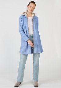 Vaaleansininen nahkatakki, jossa on vetoketju edessä, pitkät hihat ja kontrastoiva harmaa huppari. Yllä vaaleansiniset farkut ja beiget nilkkurit.