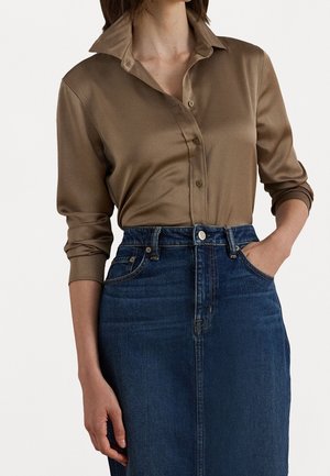 Kobieta ubrana w brązową, długorękawową koszulę z kołnierzykiem i guzikami, wsadzoną w niebieską, dżinsową spódnicę z wysokim stanem, z jedną ręką w kieszeni.