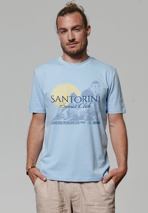 Mann mit Dutt, der ein hellblaues Santorini Sunset Club T-Shirt und beige Hosen trägt, steht mit den Händen in den Taschen vor einem einfarbigen Hintergrund.