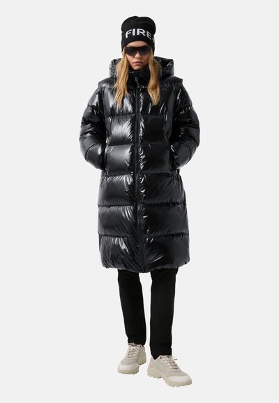 Vrouw in zwarte glanzende pufferjas met capuchon, zwarte broek, beige sneakers, zwarte muts met de tekst "FIRE" en donkere zonnebril, staande met handen in de zakken.