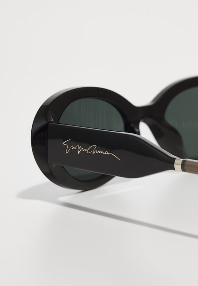 Schwarze übergroße Sonnenbrille mit grünen Gläsern. Dicke, runde Fassung und ein eleganter schwarzer Bügel mit goldfarbener Signaturakzent.