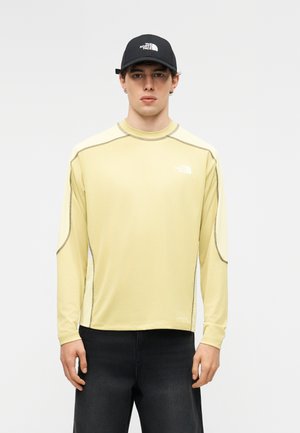 Mlad moški, oblečen v svetlo rumeno majico z dolgimi rokavi in črno kapa, obe z logotipom The North Face, stoji pred enobarvnim ozadjem.