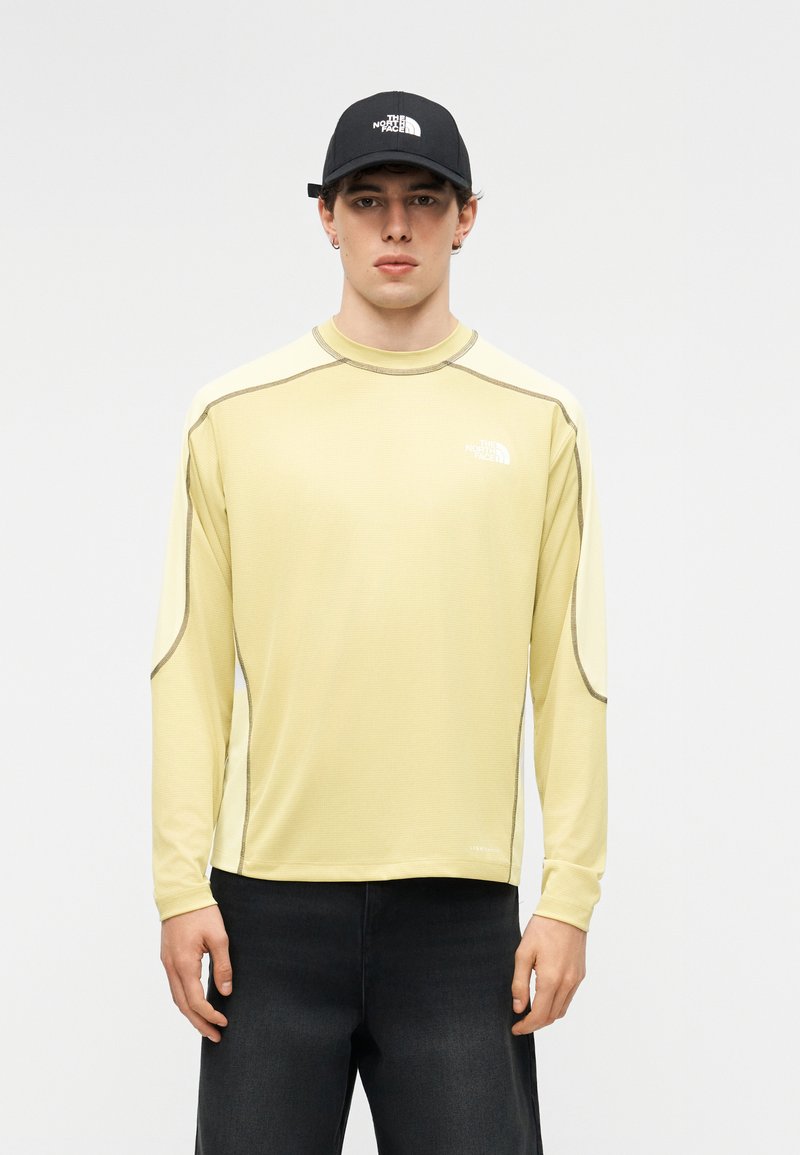 Νεαρός άνδρας που φορά ανοιχτό κίτρινο μακρυμάνικο πουκάμισο και μαύρο καπέλο, και τα δύο με το λογότυπο The North Face, στέκεται μπροστά σε απλό φόντο.