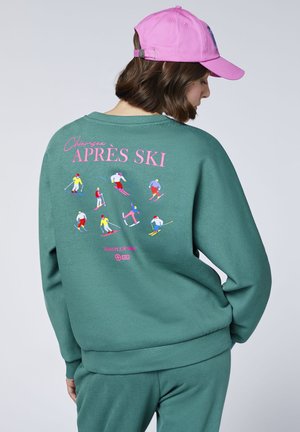 Woman wearing green sweatshirt with colorful skier illustrations and pink "APRÈS SKI" text on back, paired with pink baseball cap.