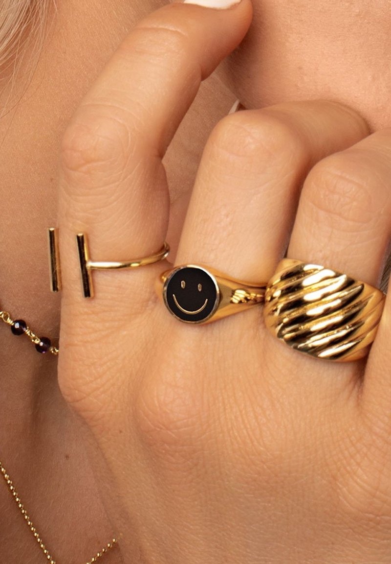 Main avec trois bagues en or : une bague ouverte, une avec un visage souriant noir et une avec un design torsadé, sur fond de peau claire.
