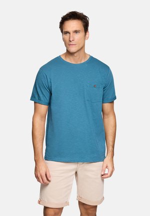 Hombre de pie de frente, lleva una camiseta de manga corta color verde azulado con un bolsillo en el pecho abotonado y pantalones cortos beige remangados.