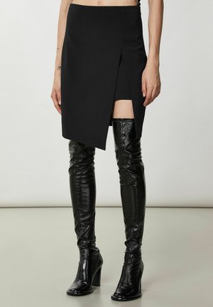 Femme portant une jupe asymétrique noire avec une fente à l'avant et des bottes hautes noires brillantes à talons, debout sur un fond clair uni.