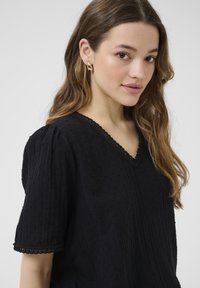 Blouse noire en tissu texturé, décolleté en V, manches courtes bouffantes et délicate dentelle. Portée avec de grandes boucles d'oreilles dorées et les cheveux lâchés.