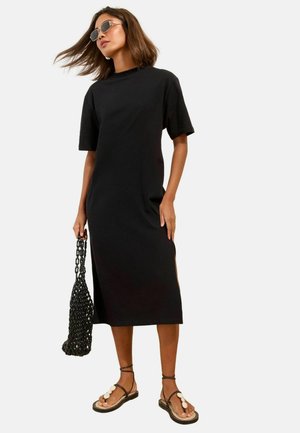 Frau trägt ein schwarzes knielanges Kleid mit kurzen Ärmeln, rundem Ausschnitt, Sonnenbrille, hält eine schwarze Netz-Tasche und trägt Muschelsandalen.