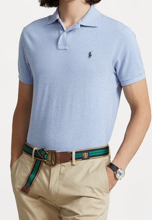 Man draagt lichtblauw poloshirt, beige broek, gestreepte riem met gouden gesp en horloge met kleurrijke band, handen in de zakken.