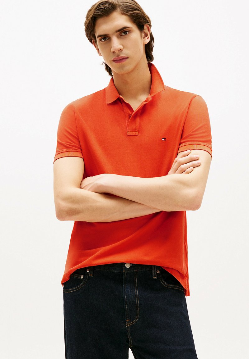 Czerwona koszulka polo wykonana z teksturowanej tkaniny, z klasycznym kołnierzykiem i krótkimi rękawami. Mały logo na lewej piersi. Połączona z ciemnoniebieskimi dżinsami.