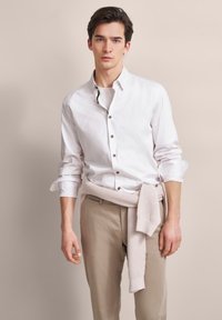 Weißes Hemd mit braunen Knöpfen, kombiniert mit beigen Hosen und einem hellbeigen Strickpullover, der um die Taille gebunden ist. Neutraler Hintergrund.