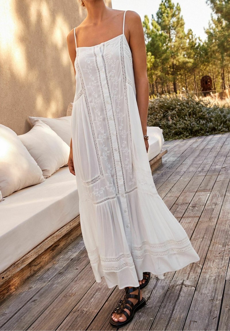 Femme portant une robe maxi blanche brodée sans manches et des sandales noires, debout sur une terrasse en bois près d’un coin salon avec coussins.