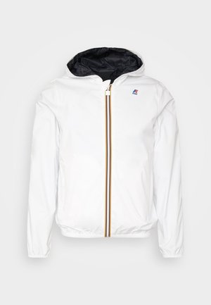 Personne debout portant une veste blanche zippée sur un polo noir, un pantalon couleur crème et des baskets blanches Adidas avec des rayures noires.