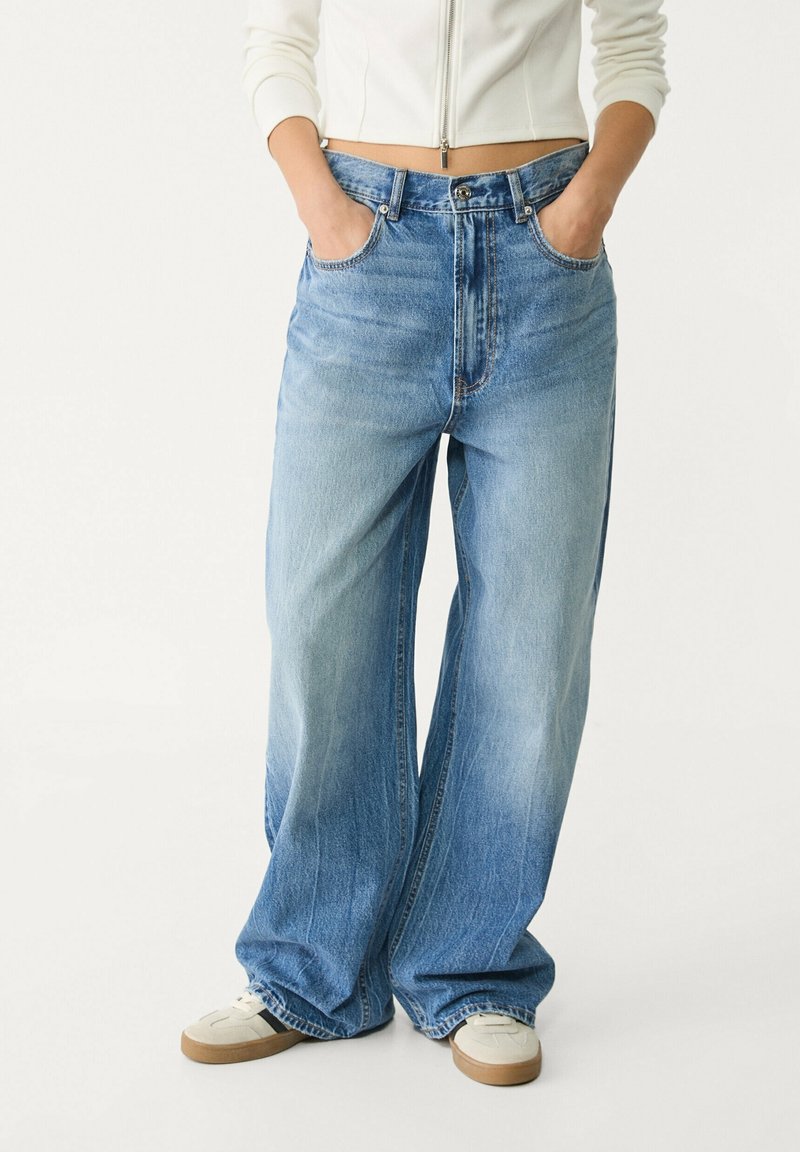 Personne portant un jean bleu clair à jambes larges avec les mains dans les poches, associé à des baskets blanches et un top blanc court zippé.