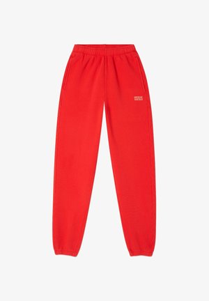 Červené sportovní kalhoty vyrobené z měkké látky, s elastickým pasem, bočními kapsami a malým potištěným logem na pravé noze.