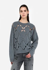 Maglione grigio lavorato a maglia con motivi floreali a traforo, scollo a girocollo e vestibilità rilassata. Abbinato a jeans grigi decorati con applicazioni.