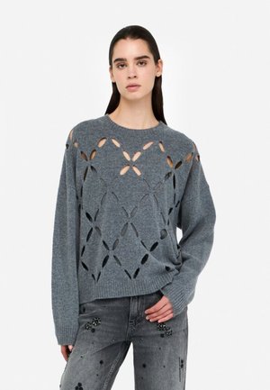 Maglione grigio lavorato a maglia con motivi floreali a traforo, scollo a girocollo e vestibilità rilassata. Abbinato a jeans grigi decorati con applicazioni.