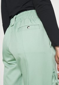 Mintgroene broek met een gerimpelde tailleband, zijzakken en een achterzak met drukknopsluiting. Gladde stoftextuur.