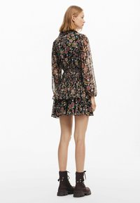 Blommönstrad klänning med svart genomskinlig tyg, långa ärmar, rynkad midja och lager av volanger i fållen, tillsammans med mörka, kraftiga sulor stövlar.