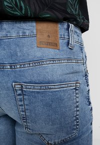 Jeans in denim chiaro con una etichetta in pelle con scritto "ONLY & SONS." Caratteristiche includono una tasca posteriore e cuciture rinforzate.