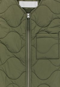 Vêtement matelassé vert olive avec une fermeture éclair argentée et une poche à rabat carrée sur le côté droit avant.