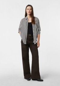 Kobieta stoi z rękami w kieszeniach, ubrana w czarno-białą poziomą pasiastą koszulę, czarną bluzkę, ciemne szerokie spodnie i czarne buty na białym tle.