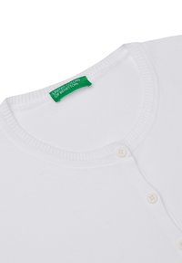 Bílý bavlněný cardigan s kulatým výstřihem, který má žebrovaný límec, dva bílé knoflíky a zelenou etiketu s nápisem "United Colors of Benetton."