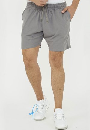 Mann trägt graue elastische Shorts mit Kordelzug in der Taille und weiße Sneaker mit blauem Logo, steht mit einer Hand in der Tasche.
