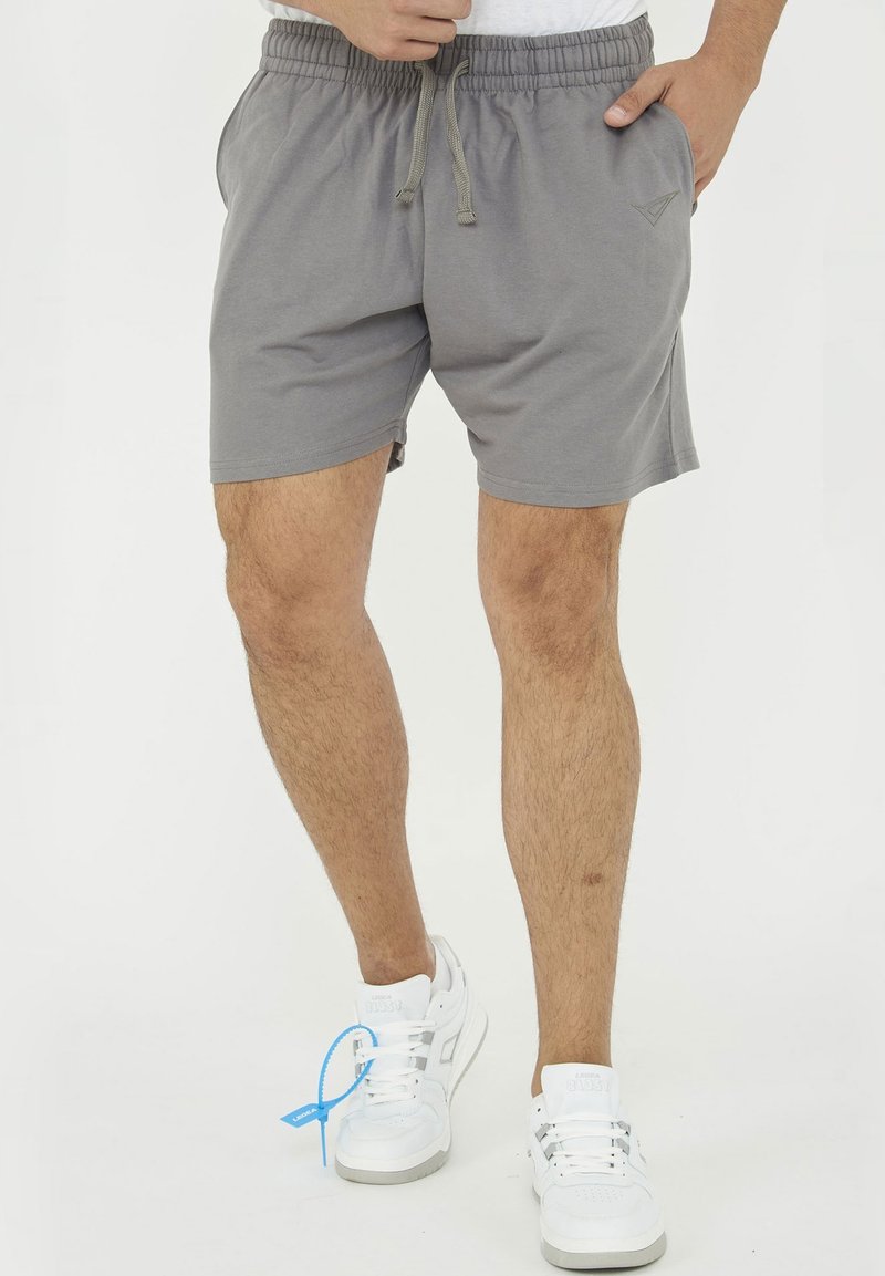 Homme portant un short gris à taille élastique avec cordon et des baskets blanches avec une étiquette bleue, debout avec une main dans la poche.