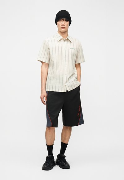 Νεαρός άνδρας με μαύρο σκουφάκι, μπεζ κοντομάνικο πουκάμισο με ρίγες, μαύρες και γκρι βερμούδες με κόκκινες λεπτομέρειες, μαύρες κάλτσες και μαύρα αθλητικά παπούτσια.