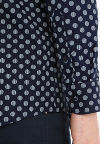 Marineblauw overhemd met een patroon van witte gestreepte polkadots, met een geruite mouw en een gladde textuur. Getailleerd ontwerp met een lichte taps toelopende pasvorm.