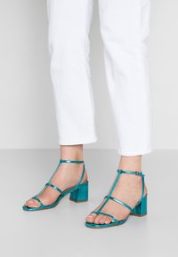 Tyrkisk blå hæle sandaler med metallisk finish, der har tynde stropper og en blokhælsdesign. Udsatte tæer med ankelstropper.