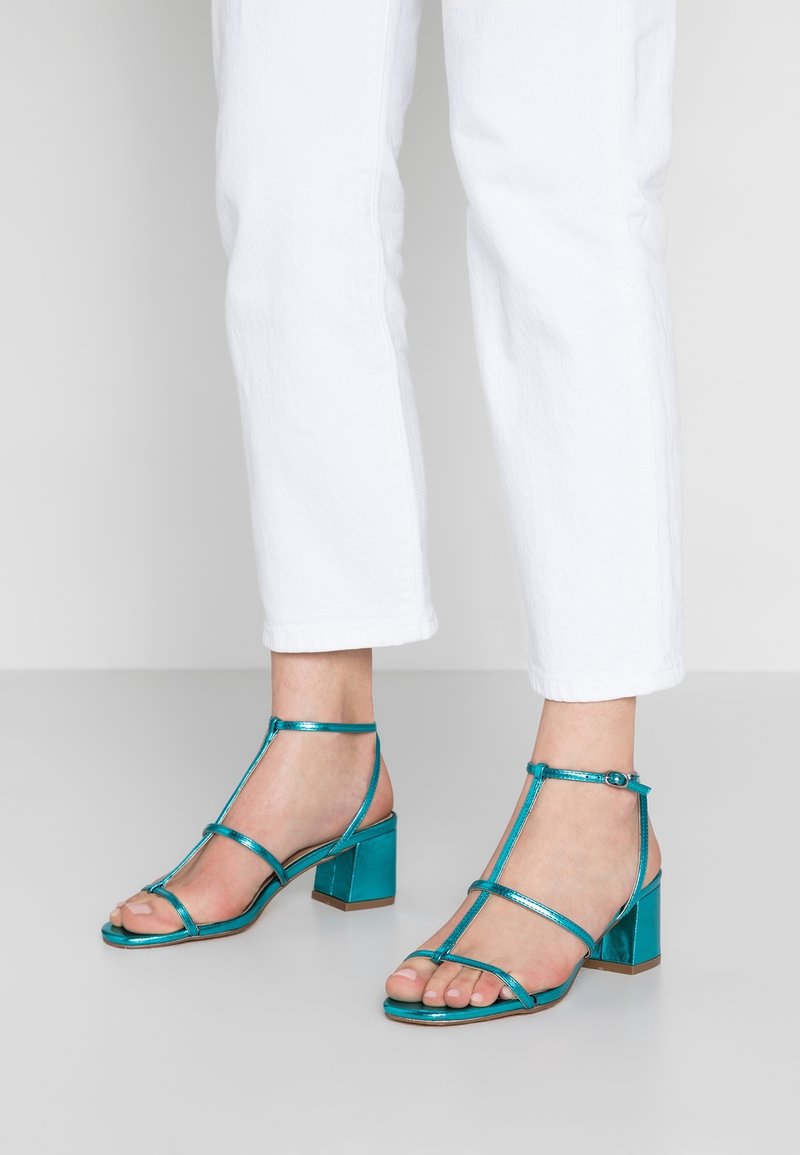Tyrkisk blå hæle sandaler med metallisk finish, der har tynde stropper og en blokhælsdesign. Udsatte tæer med ankelstropper.
