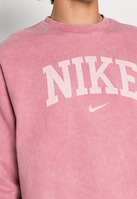 Sweatshirt rose en tissu doux. Doté d'un grand logo "NIKE" blanc sur la poitrine et d'un petit logo swoosh Nike en dessous. Encolure ronde.