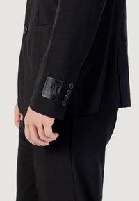 Persona che indossa un completo nero con quattro bottoni sulla manica e un patch con logo "AX" vicino al polsino.