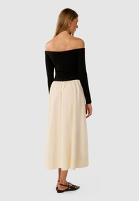 Frau mit dem Rücken zur Kamera, die ein schwarzes, schulterfreies Langarmoberteil, einen cremefarbenen Midirock und schwarze spitze Ballerinas mit Knöchelriemen trägt.