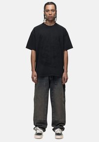 T-shirt nera oversize con un design grafico testurizzato, abbinata a jeans in denim scuro a vestibilità ampia e sneakers bianche con dettagli neri.