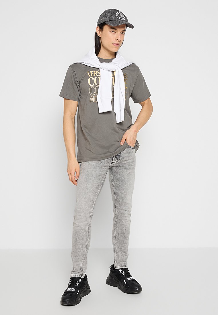 Maglietta grigia con stampa dorata; maglione bianco appoggiato casualmente sulle spalle; jeans grigi chiari; sneaker nere; cappellino grigio con logo.