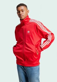 Ung mann ikledd en rød Adidas treningsjakke med hvite striper og blå jeans, stående med hendene i lommene mot en ensfarget bakgrunn.