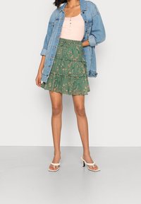 Falda mini floral verde con volantes, combinada con una camiseta ajustada de color rosa claro. Una chaqueta de denim azul y sandalias de tacón color crema completan el look.