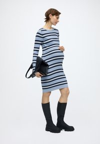 Εφαρμοστό, ριμπ φόρεμα εγκυμοσύνης με μπλε και ναυτικά ριγέ. Συνδυάζεται με μαύρες μπότες που φτάνουν μέχρι το γόνατο. Κρατά μια μαύρη δερμάτινη τσάντα ώμου.