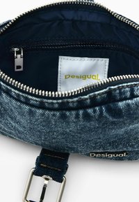 Jeans-Umhängetasche mit silbernem Reißverschluss, Metallverschluss und Innentasche, die ein weißes Etikett mit "Desigual"-Logo in Gelb und schwarzer Naht zeigt.