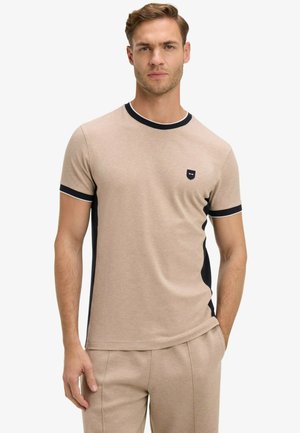 Homme aux cheveux châtains courts portant une chemise à manches courtes beige et noire ainsi qu'un pantalon beige assorti, debout avec une main dans la poche.