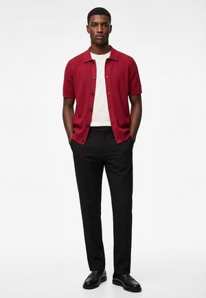 Uomo in piedi con le mani in tasca, indossa una camicia rossa a maniche corte con bottoni, una maglietta bianca, pantaloni neri e scarpe eleganti nere.