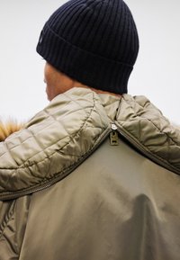 Hombre que lleva un gorro de lana negro y una chaqueta verde oliva con un cuello acolchado con cremallera, visto desde atrás.