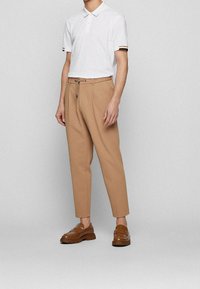 T-shirt polo blanc avec des poignets rayés, associé à un pantalon beige ajusté et des mocassins marron, doté d'une taille à cordon. Texture lisse.