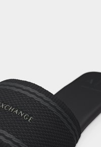 Sandale à glissière noire texturée avec une bride surélevée comportant la marque "EXCHANGE" en relief et des détails de couture décoratifs.